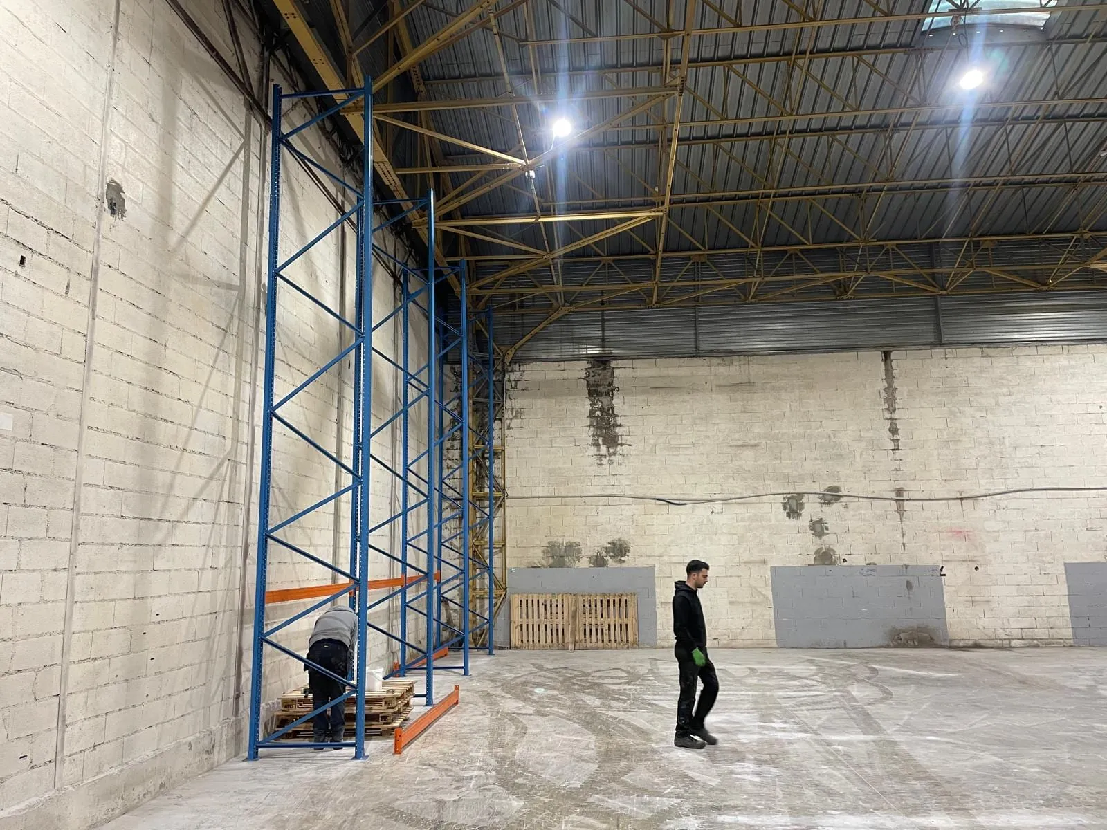 De eerste blauwe jukken van een nieuwe palletstelling tegen de wand van een lege hal, met een monteur aan het werk aan de voet