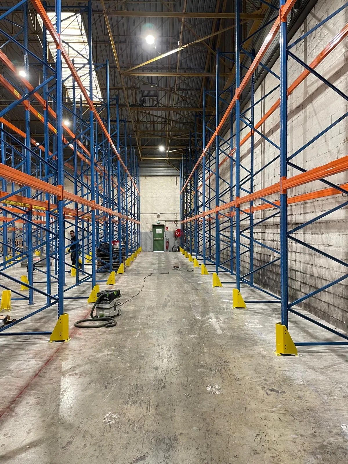 Gang tussen twee rijen blauwe palletstellingen met oranje liggers, met gele aanrijdbeveiliging aan de voet van elke staander