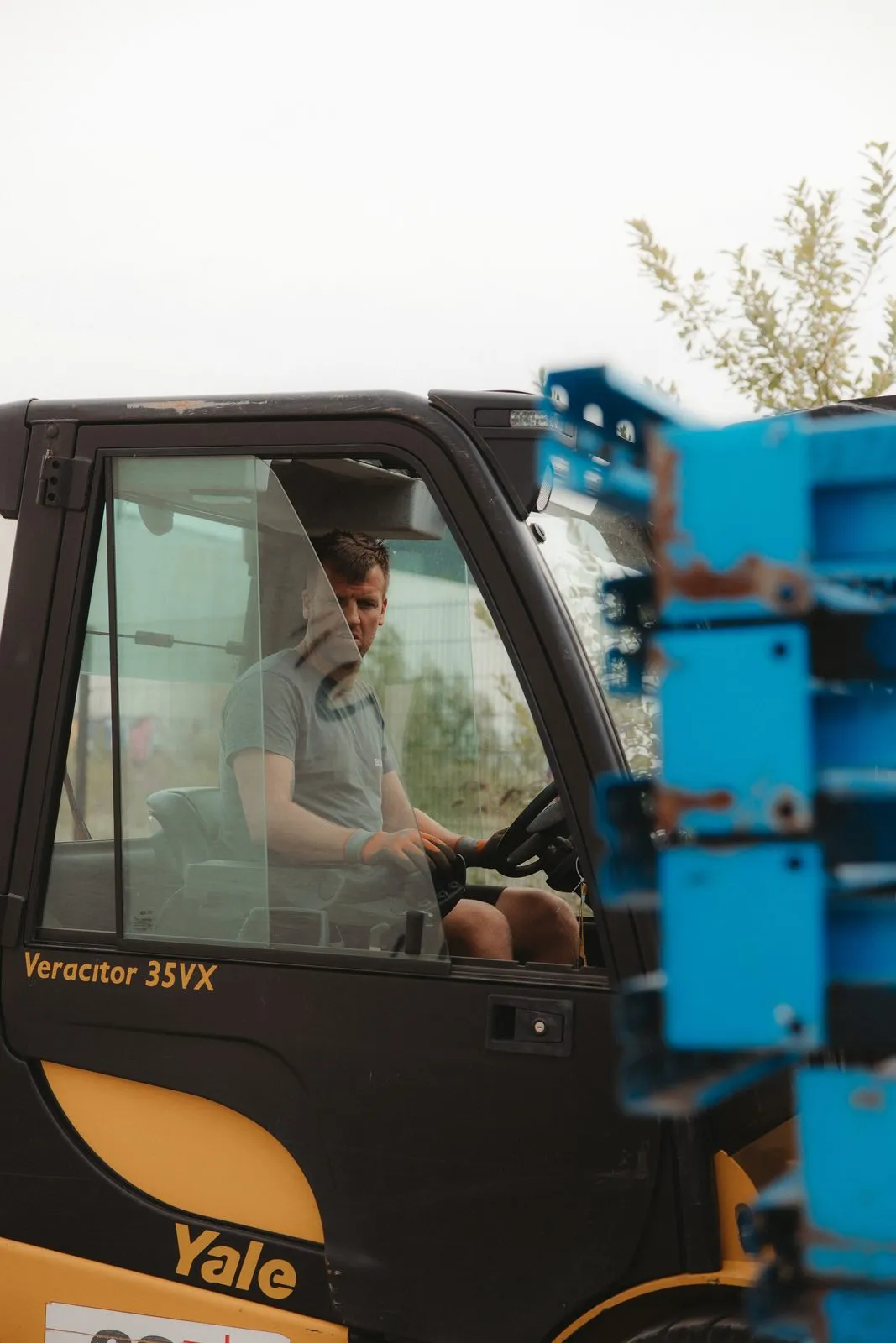 Bram achter het stuur van een gele heftruck, met blauwe staanders voor de cabine