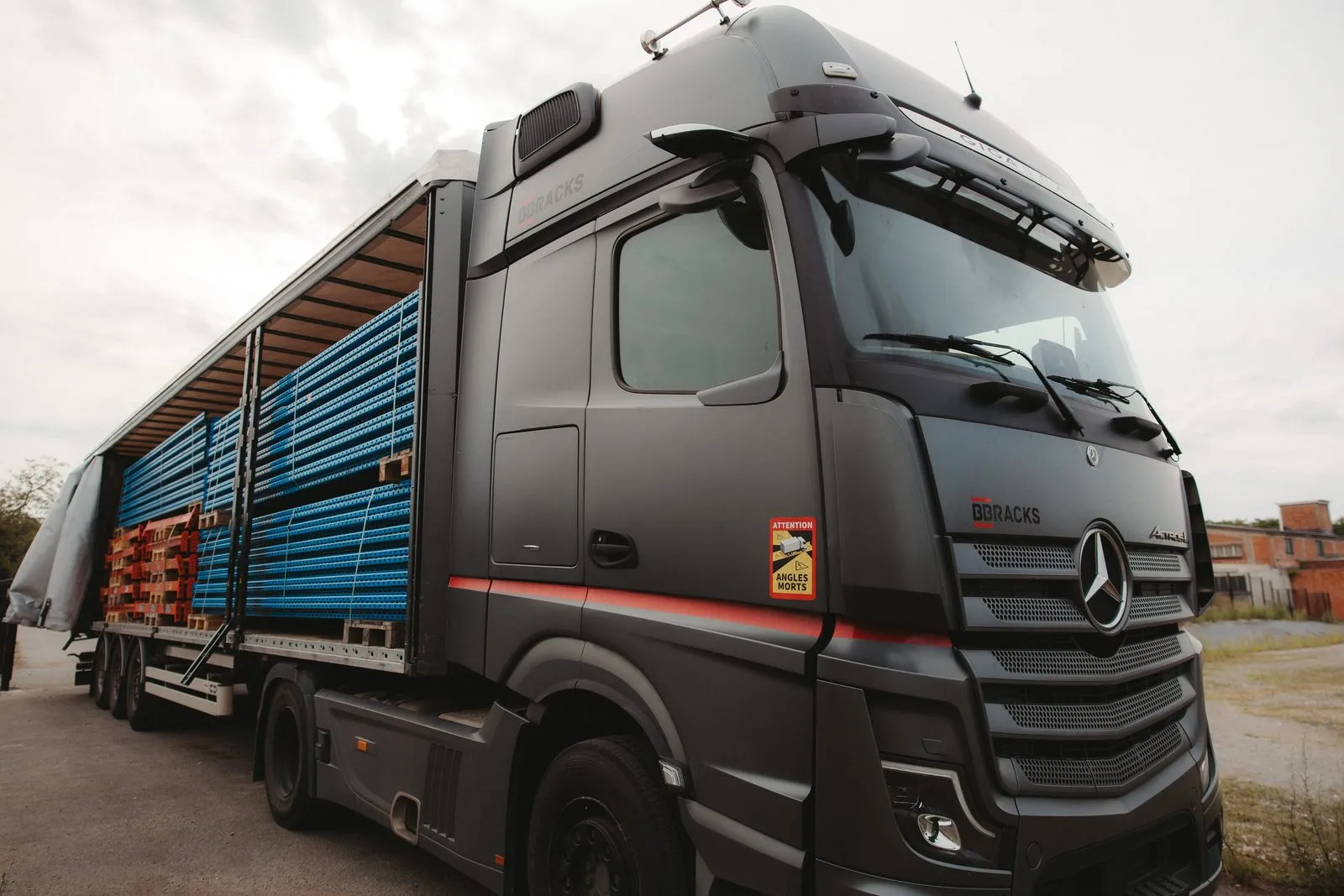 Zwarte vrachtwagen met het BB Racks-logo op de cabine, de oplegger open en geladen met blauwe staanders en oranje liggers