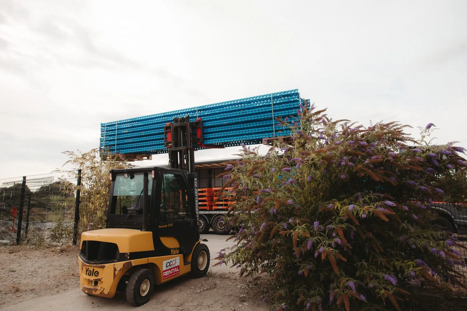 Een gele heftruck tilt een bundel blauwe staanders over een bloeiende struik