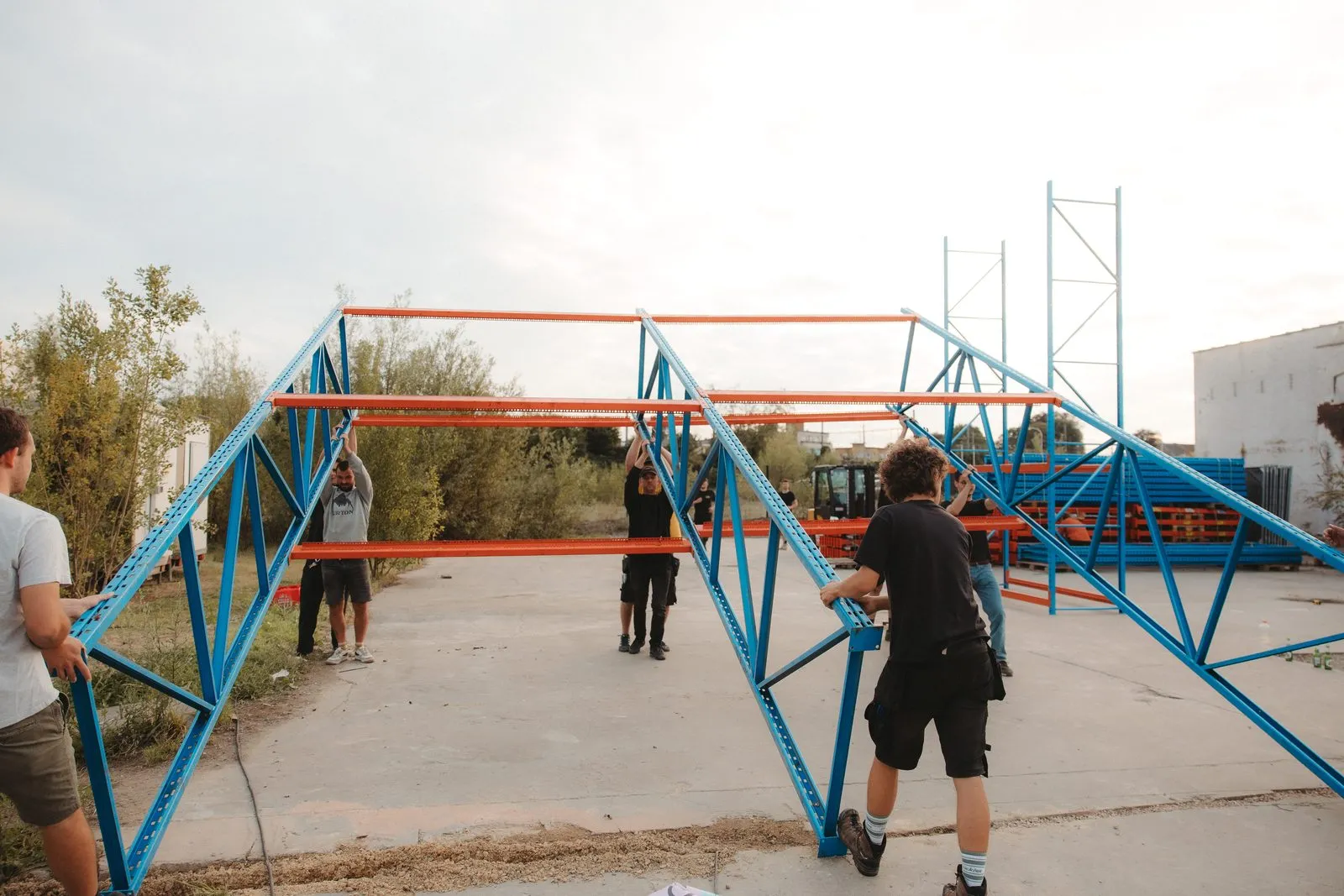 Een groep mensen zet twee blauwe rekken met oranje liggers rechtop op een betonnen terrein