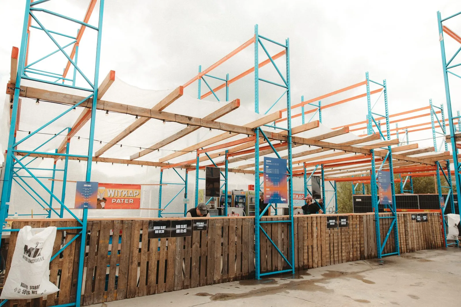 Een bar onder een overkapping van blauwe staanders, oranje liggers, houten balken en een wit net, met een toog van paletten