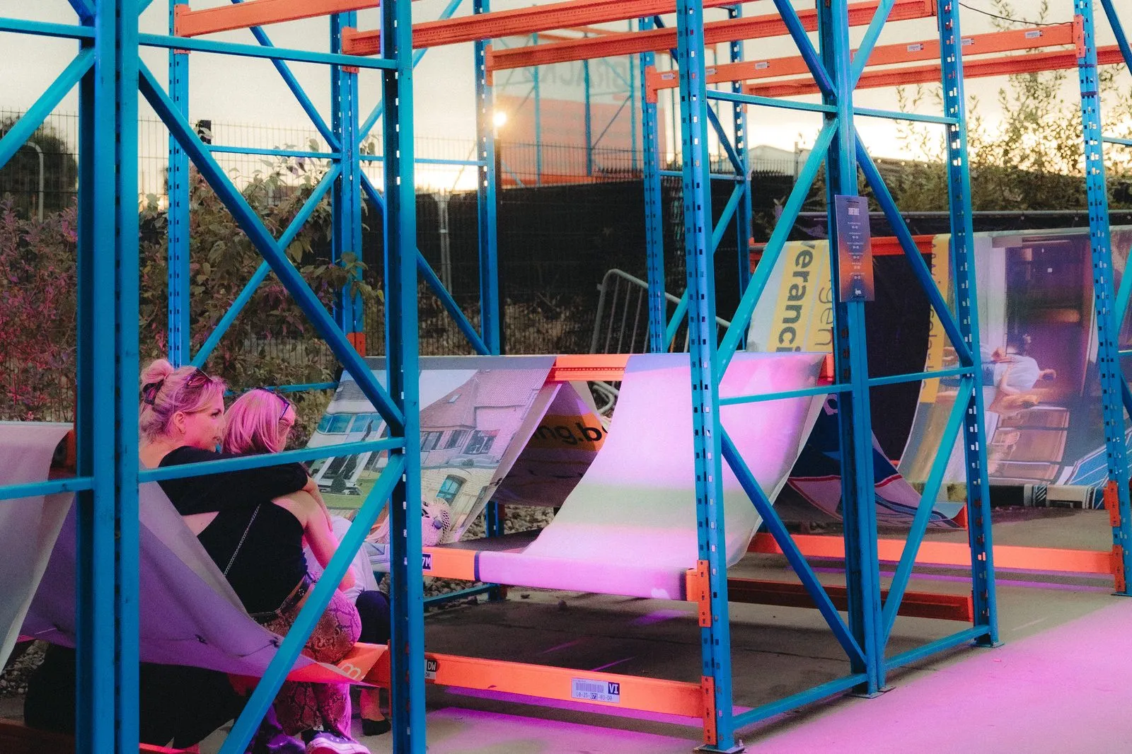 Ligzetels van doek hangen tussen de onderste oranje liggers van blauwe rekken, in roze avondlicht