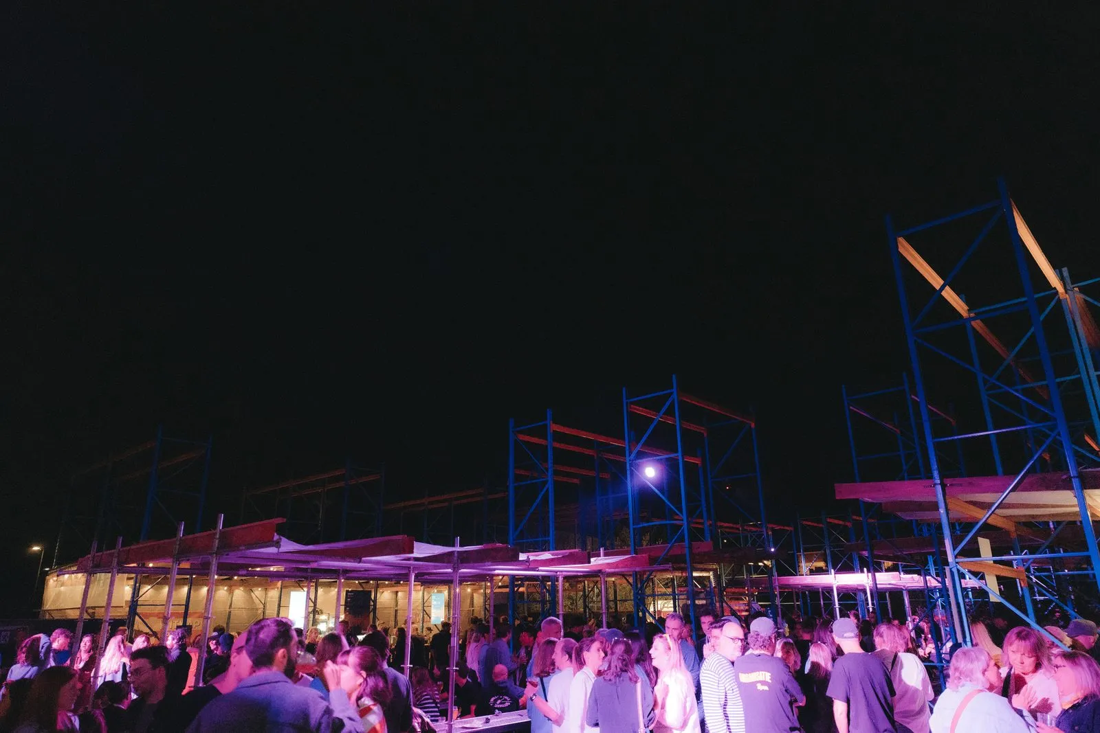 Het festivalterrein bij nacht, met verlichte overkappingen en blauwe rekken boven de bezoekers